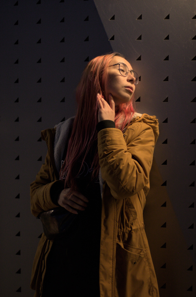 Woman with pink hair and eyes closed leaning on a wall metal with a pattern, under soft warm street light