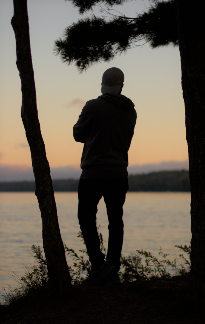 Man in a white cap looking away on a sunset next next to a lake, under a tree